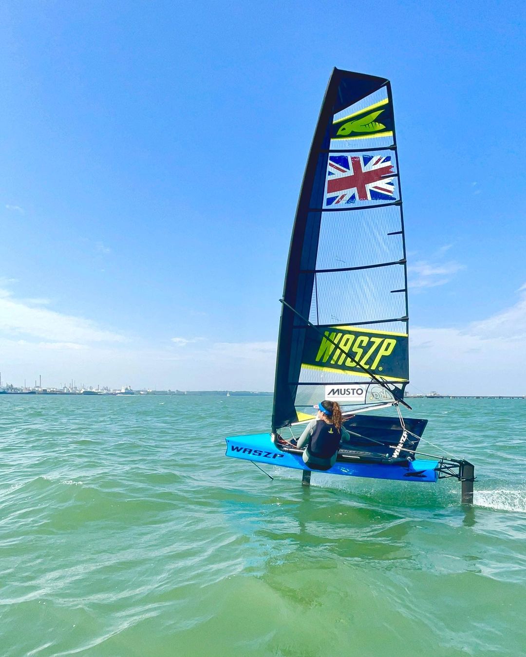 Waszp foiling dinghy with a Union Jack sail lifting clear of turquoise shallow water