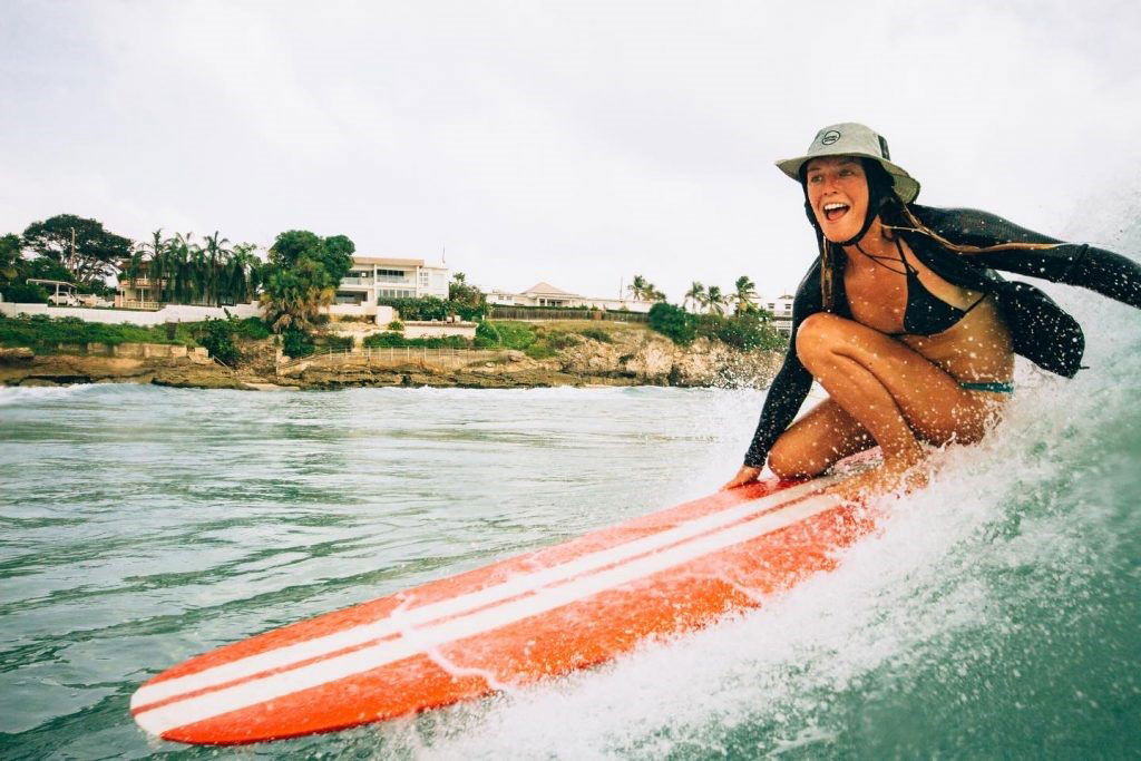 Smiling surfer in a bucket hat riding an orange longboard along a clean tropical wave