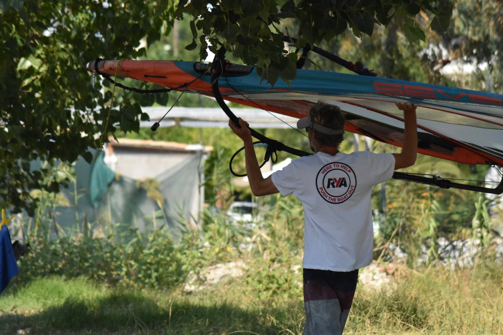 Windsurfer carrying a rigged sail on his shoulder through a garden towards the water