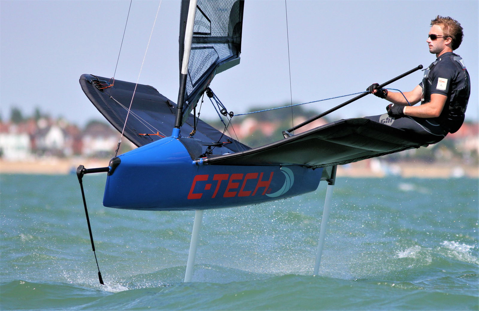 Sailor sitting out on the wing of a blue foiling Moth lifted clear of the water