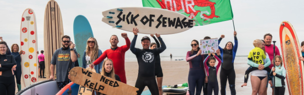 Beach protest with surfers holding boards reading Sick of Sewage and We Need Kelp, and a Welsh flag