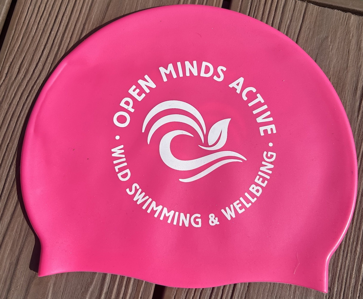 Pink silicone swim cap printed Open Minds Active Wild Swimming and Wellbeing, lying on decking