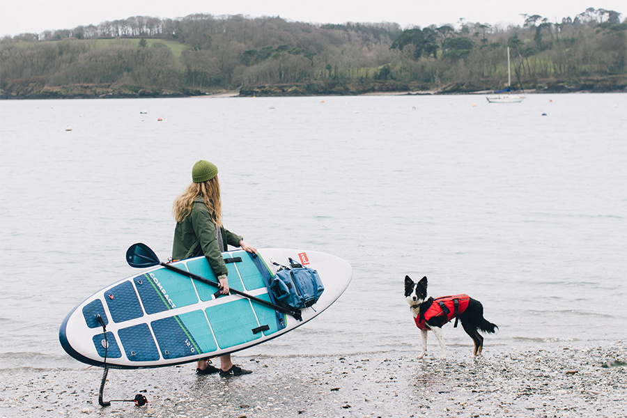 Woman carrying a teal inflatable paddleboard down a beach with a dog in a red buoyancy aid